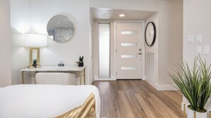 Een hotelkamer met een bed, een bureau met lamp, een ronde spiegel, een deur met raam en een pot met plant.