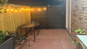 Un área de patio al aire libre con cerca de madera, una mesa con sillas y luces colgantes.