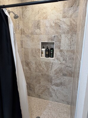 A shower area with beige tiles, a showerhead, a towel rack, and a shelf with three bottles. - Peaceful & Private Studio #2 (Brownsville)