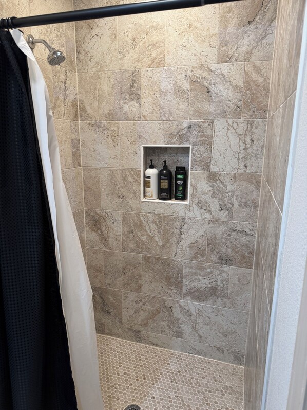 A shower area with beige tiles, a showerhead, a towel rack, and a shelf with three bottles.