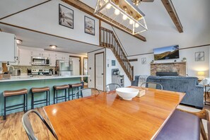 A spacious kitchen with modern appliances, a dining area with a large wooden table, and a cozy living room with a grey sofa and fireplace.