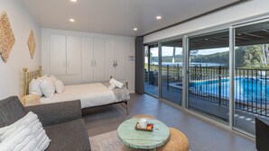 A bedroom with a bed, a sofa, a small table, and a view of a swimming pool through large sliding doors. - Tambo Lindo- Meaning beautiful inn in Quechua the Inca language of Perú (Smiths Lake)