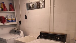 A laundry area with two white washers and dryers, a shelf with cleaning supplies, and a 'Laundry' sign on the wall.