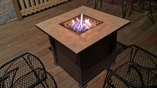 A square fire pit table with two metal chairs on a wooden deck.