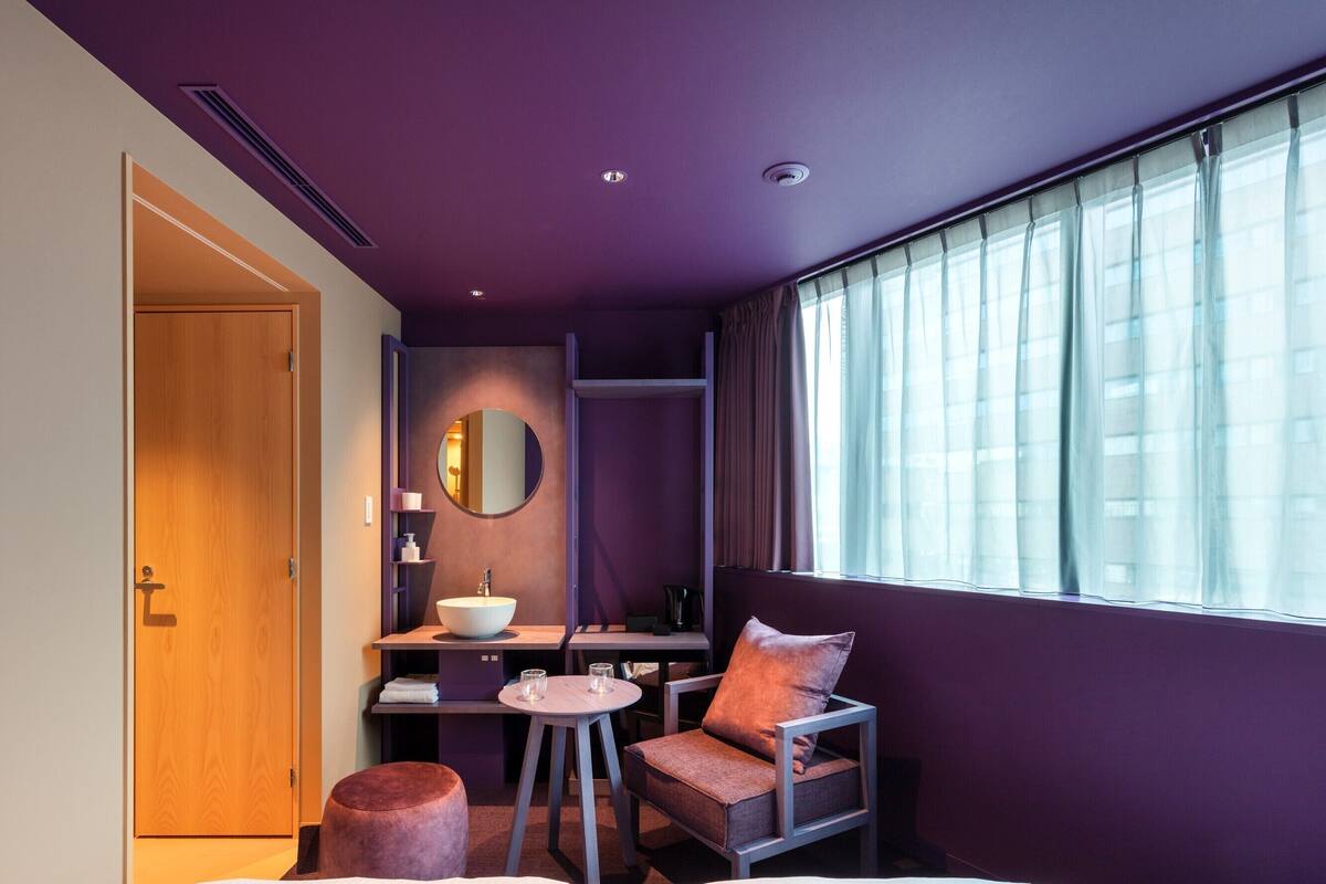 A modern hotel room with a large bed, a small round table, a chair, a sofa, a bathroom with a sink and mirror, and a window with curtains.
