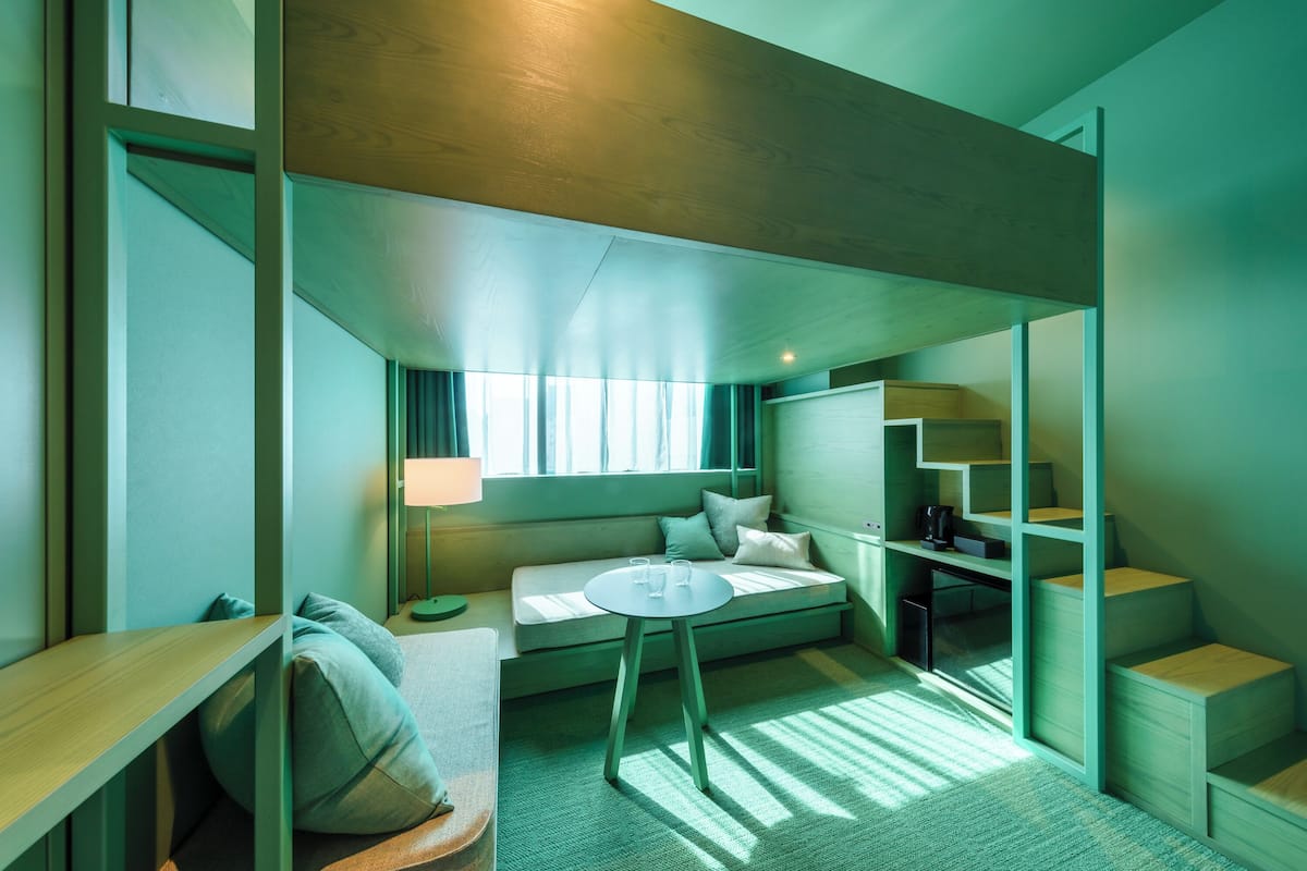 A modern hotel room with a built-in sofa, a small round table, a desk, and a staircase.
