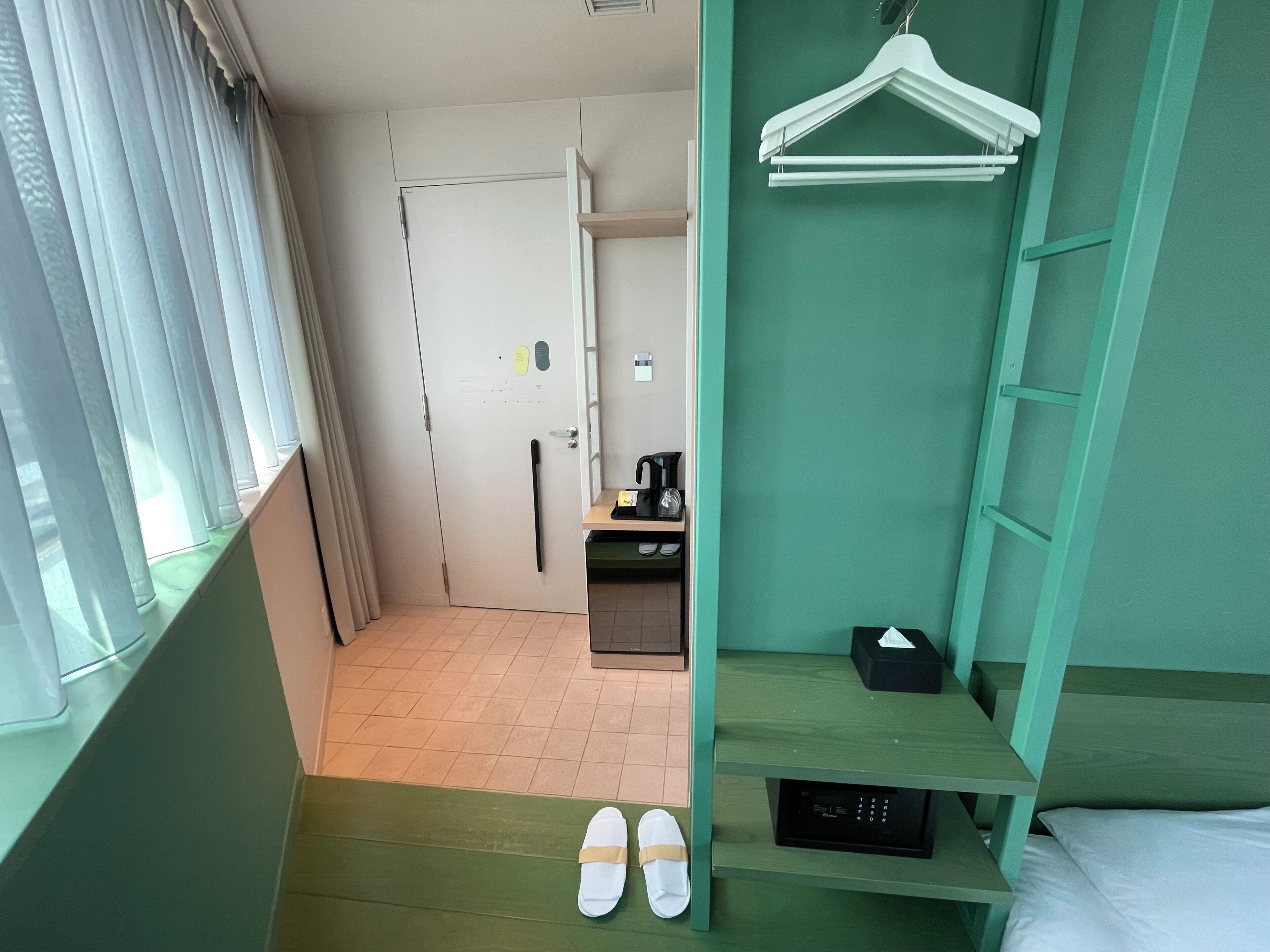 A hotel room with a bed, a desk with a coffee maker, a hanging clothes rack, and a pair of slippers on the floor.