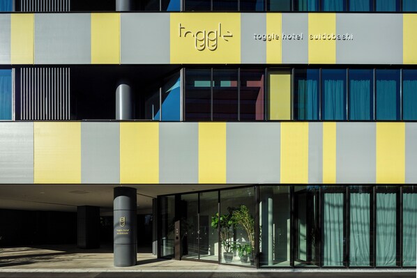 A modern hotel with a yellow and gray facade, large glass windows, and a visible entrance area with potted plants. - toggle hotel suidobashi TOKYO (Tokyo)