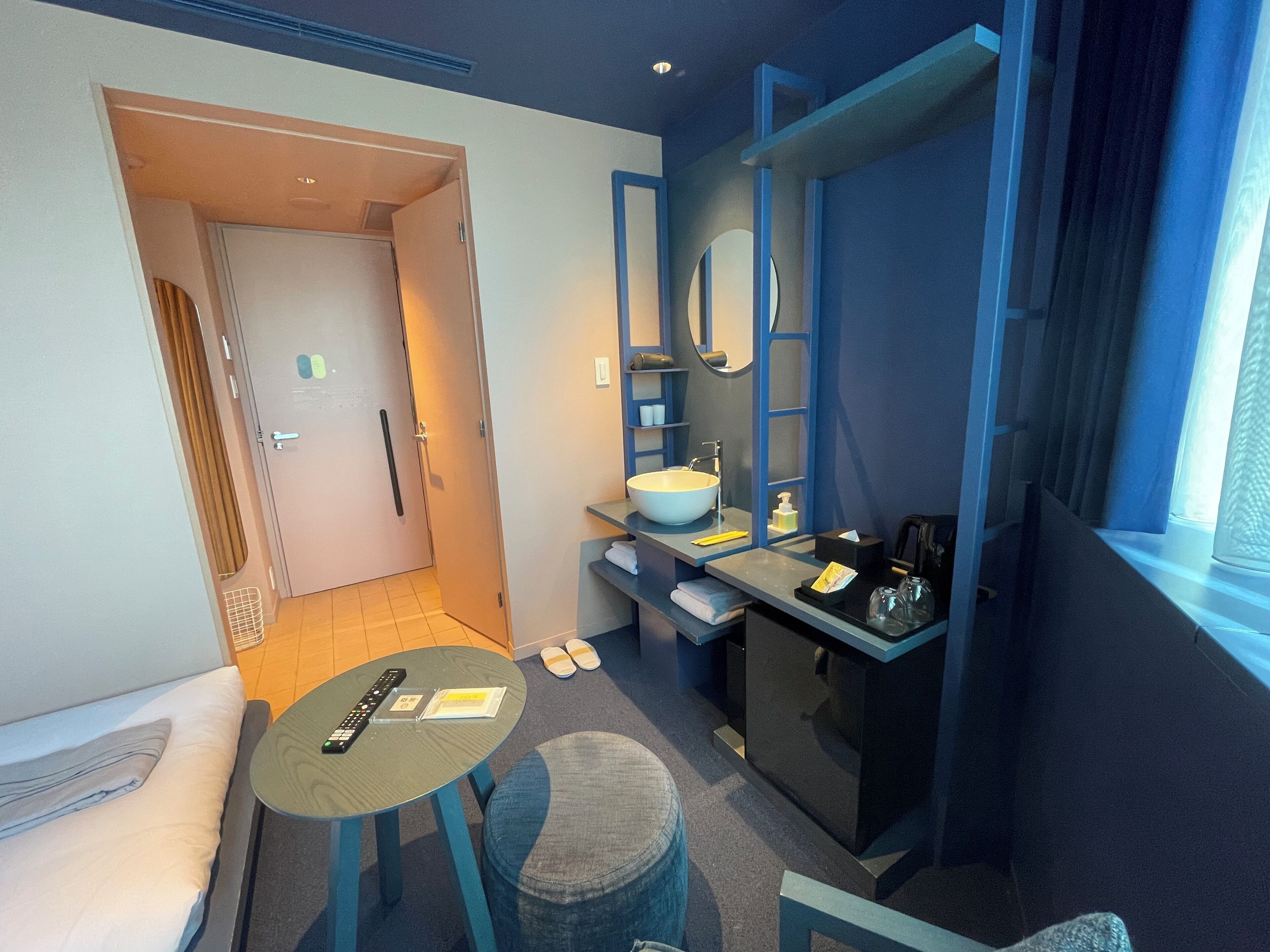 A compact hotel room with a bed, a small table, a round ottoman, a bathroom with a sink and mirror, and a window with curtains.