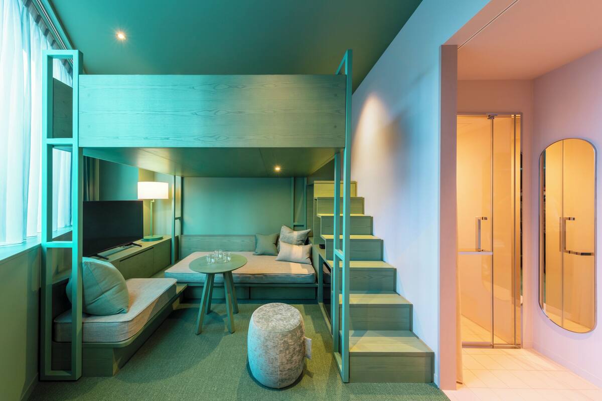 A compact hotel room with a sofa, a small table, a TV, and a stone ottoman. There is a staircase leading to a shower area.