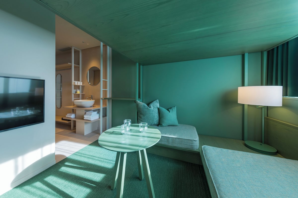 A modern hotel room with a flat-screen TV, a round table with glasses, a sofa with pillows, a lamp, and a bathroom visible through an open door.