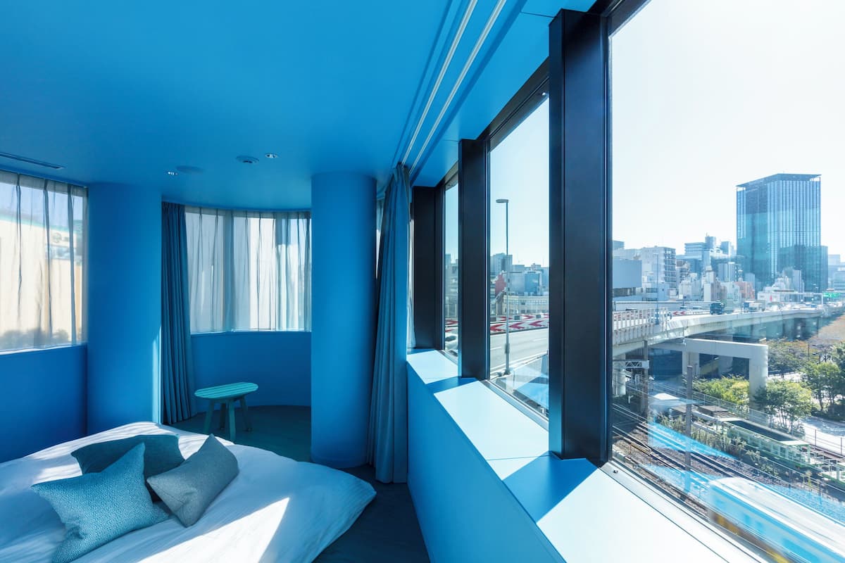 A modern hotel room with a large bed, a small table, and a city view through large windows.