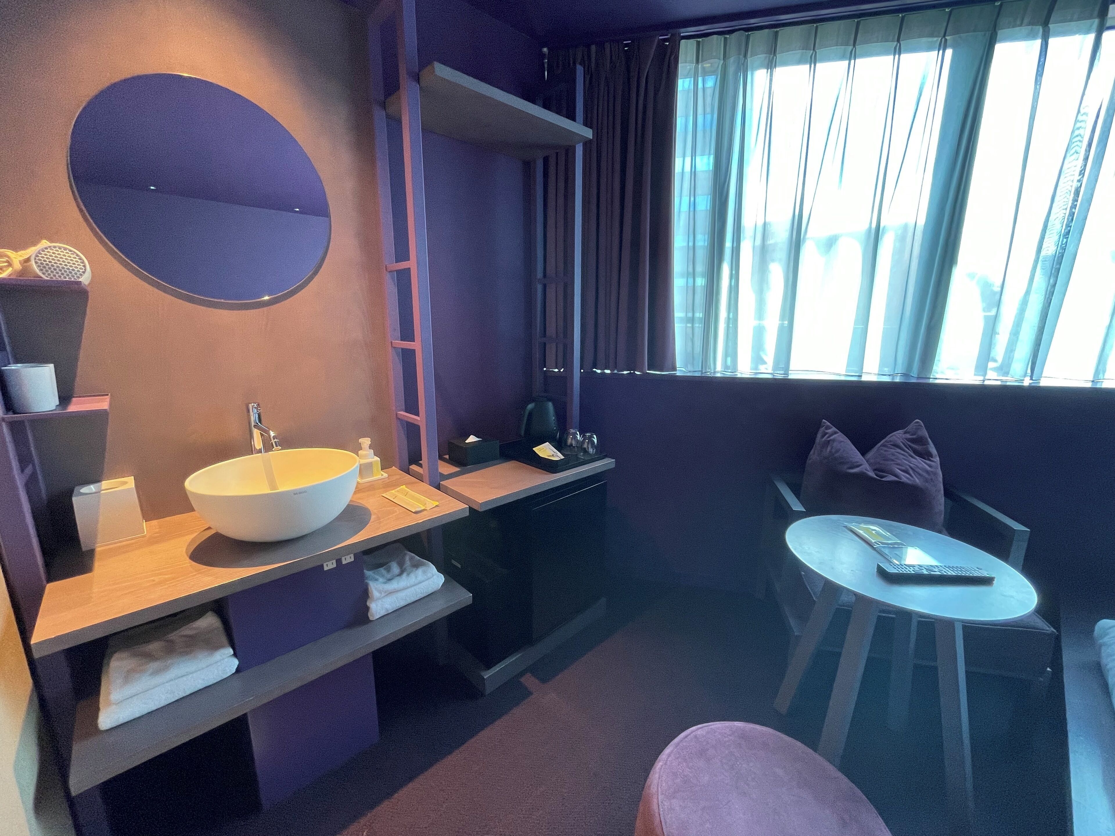 A modern bathroom with a large round mirror, a white sink, and a glass-enclosed shower. There is a small round table with a chair and a purple sofa in the adjacent living area.