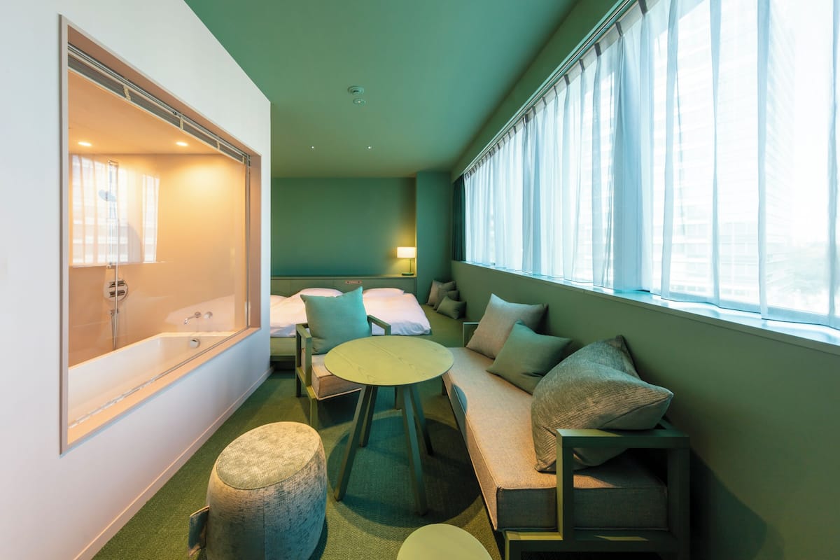 A modern hotel room with a large window, a sofa, a round table, and a bathtub.