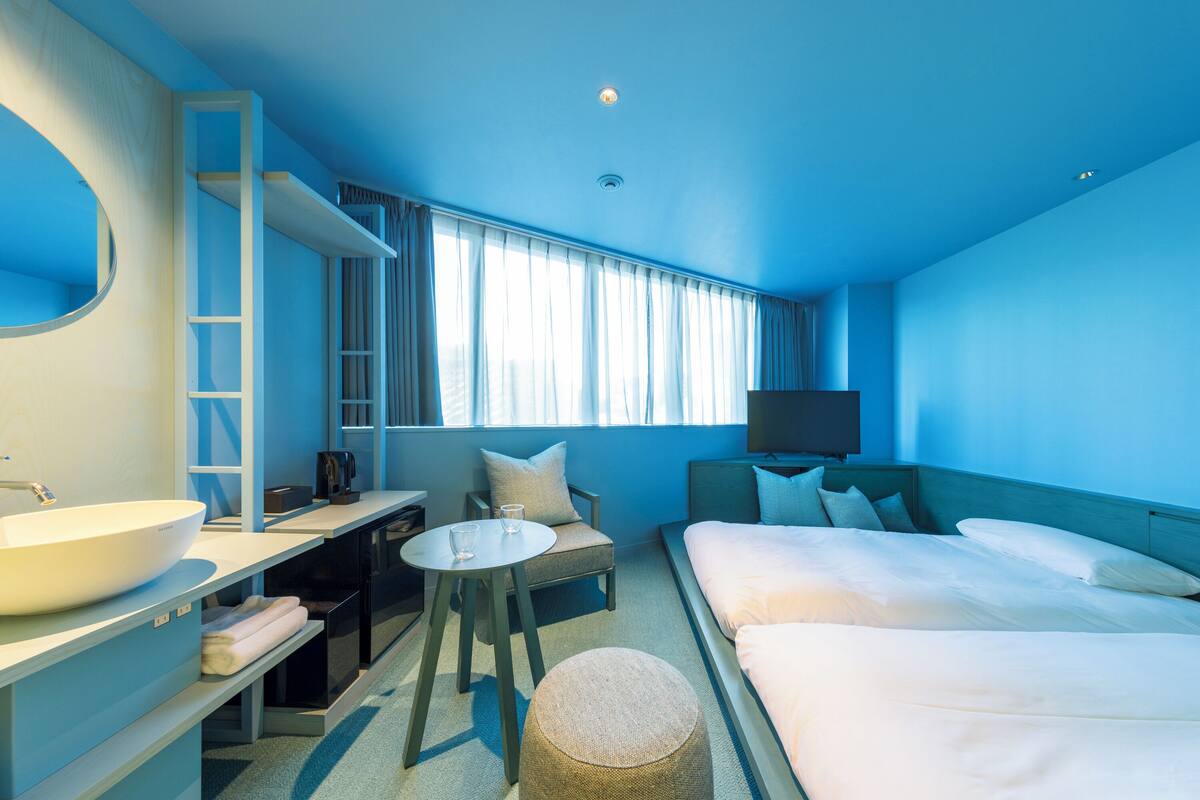 A hotel room with a large bed, a desk with a sink, a round ottoman, and a TV on the wall.