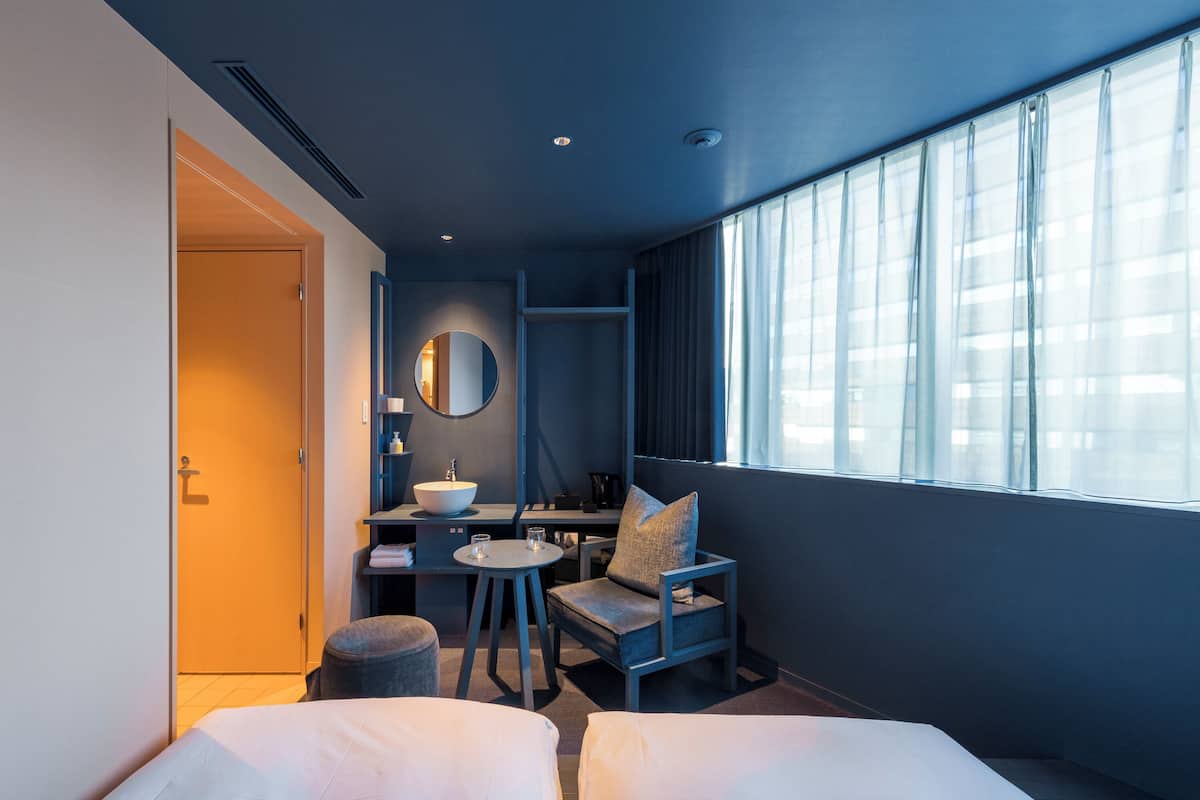A modern hotel room with a bed, a small desk with a chair, a round mirror, and a window with curtains.