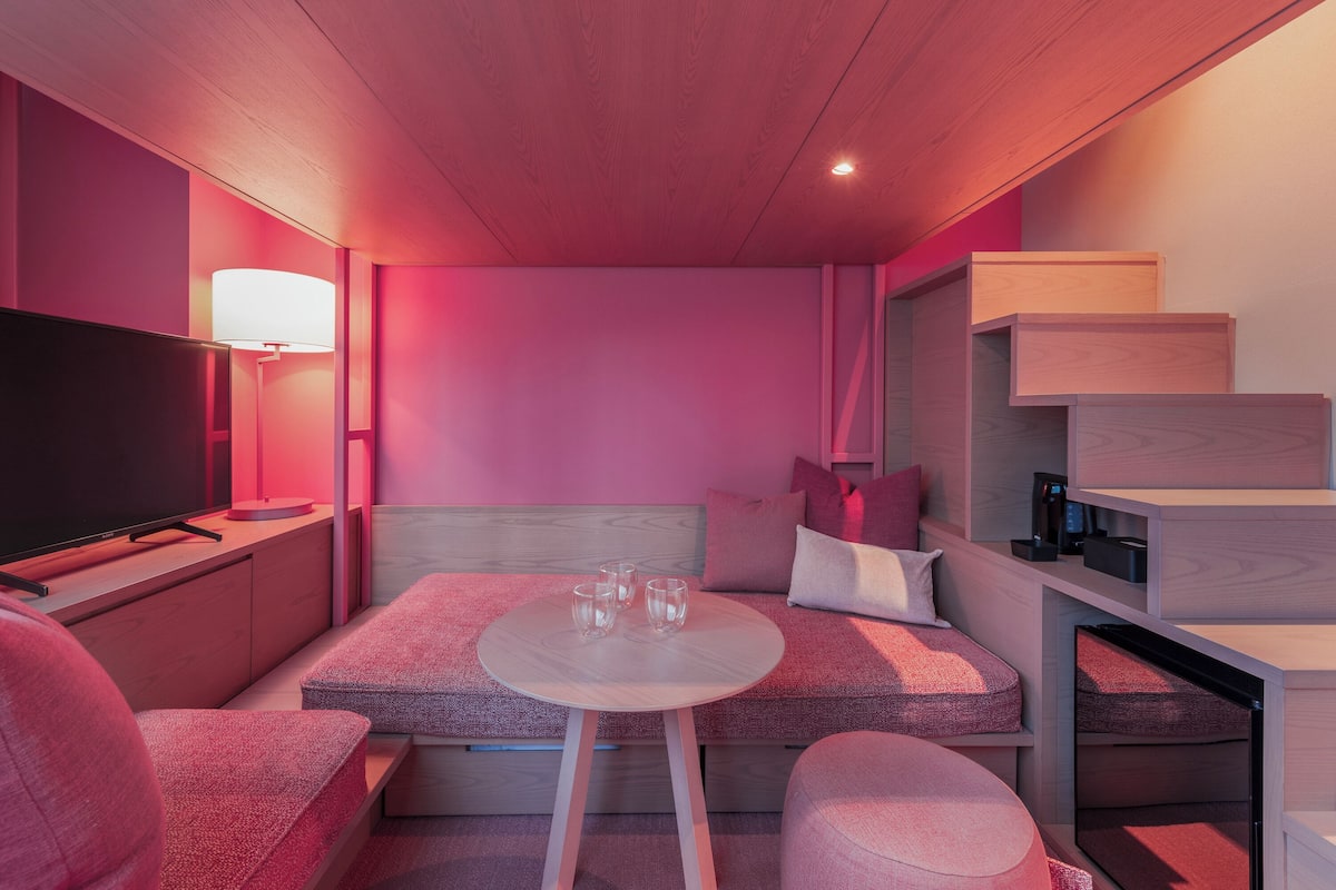 A modern hotel room with a TV, a round table with glasses, a sofa, and a staircase.