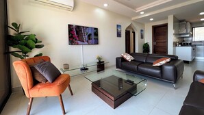 A modern living room with a glass coffee table, a sofa, an orange armchair, and a TV mounted on the wall.