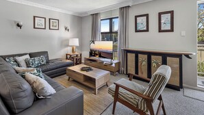 Un salon comprenant un canapé, un fauteuil, une table basse et un téléviseur. Les murs sont ornés de tableaux encadrés et il y a une fenêtre avec des rideaux.