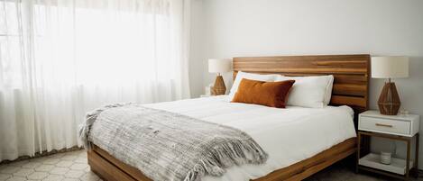 Un letto rifatto con cura, con testiera in legno, biancheria bianca e una coperta grigia. Sono presenti due comodini con lampade e una piccola mensola.