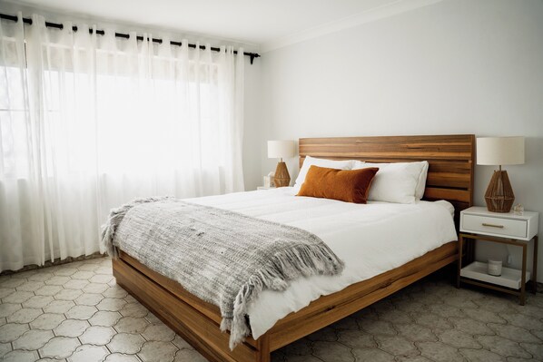 A neatly made bed with a wooden headboard, white bedding, and a gray throw blanket. There are two bedside tables with lamps and a small shelf. - Family Getaway With Ocean Views (Gerroa)