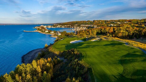 A coastal golf course with a view of the sea, a sandy beach, and a forested area.