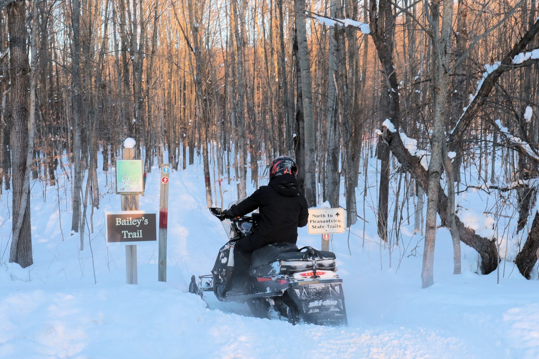 A snowmobile on a trail with signs indicating directions to Bailey's Trail and Pleasantview State Trails.