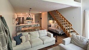 A spacious living area with a high ceiling, a wooden staircase, and a kitchen in the background.
