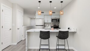 Una cocina moderna con armarios blancos, un refrigerador de acero inoxidable, un microondas e isla blanca con dos taburetes de bar.