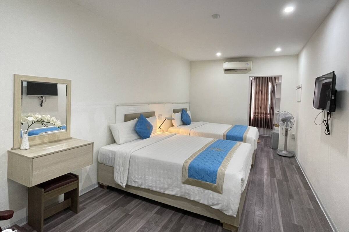 A hotel room with two beds, a bedside table with a lamp, an air conditioning unit, a TV, and a fan.