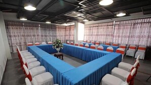 Un salón de conferencias con una larga mesa rodeada de sillas, una mesa pequeña con un florero en el centro y cortinas en las ventanas.