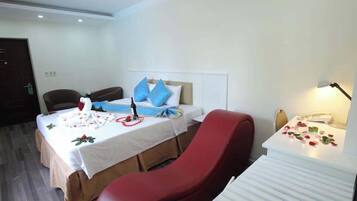 A hotel room with a bed, a red chair, a small table, and a desk with a lamp.