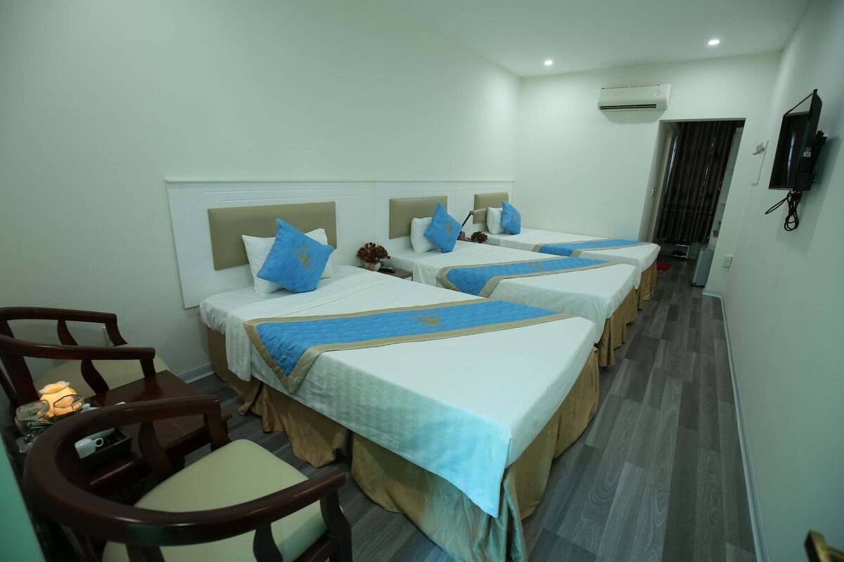 A hotel room with three beds, each with a blue headboard and matching pillow. There is a wooden chair and a small table with a lamp.