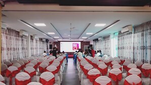 Un salón de conferencias con filas de sillas, un escenario con una pantalla y un ventilador de techo.