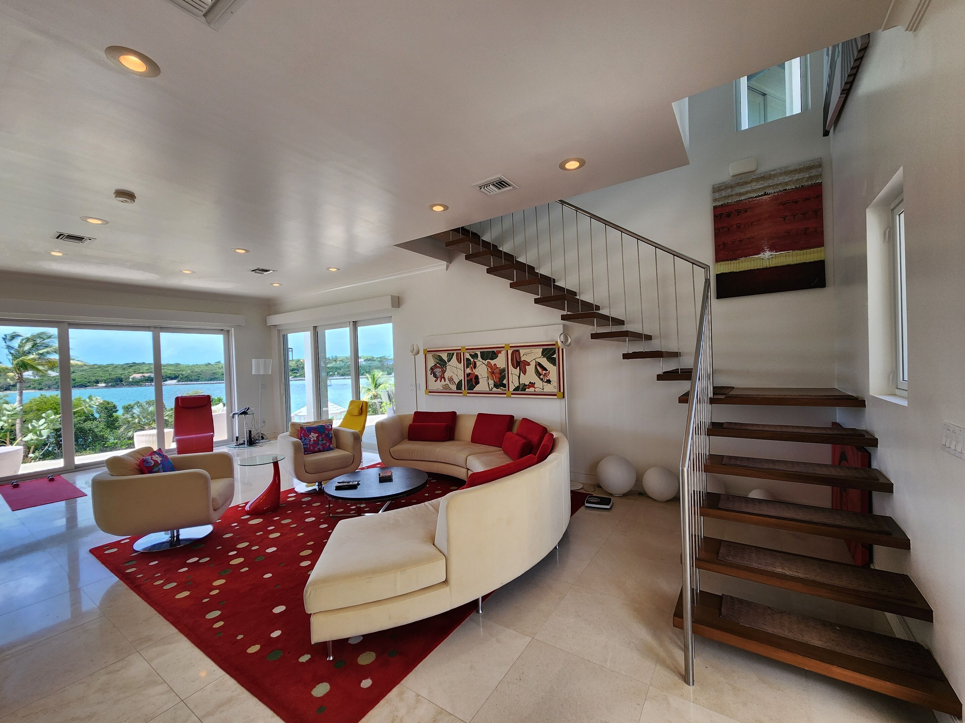 Een moderne woonkamer met een gebogen witte bank, rode kussens, een rood tapijt met patroon en uitzicht op zee door grote ramen.