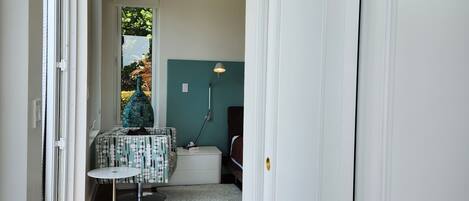 A hallway with wooden flooring, a white door, and a view into a room with a bed and a lamp.