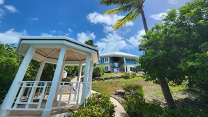 A gazebo with a wooden floor and white railings, a palm tree, and a house with a blue roof and white walls. - "Gorgeous, 4-bedroom, 4.5-bathroom home with Beautiful Water and Sunset Views!" (George Town, Great Exuma)