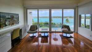 A spacious room with a large window overlooking a scenic ocean view, two modern chairs, a wooden floor, and a painting on the wall. - "Gorgeous, 4-bedroom, 4.5-bathroom home with Beautiful Water and Sunset Views!" (George Town, Great Exuma)