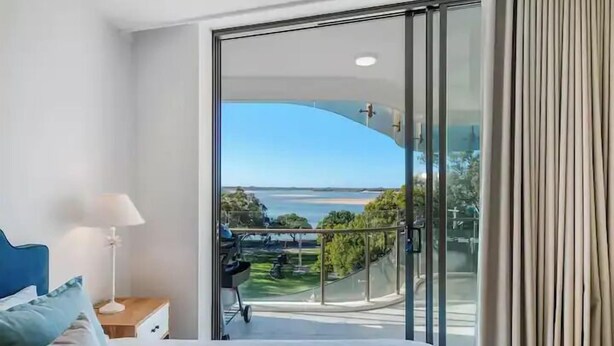 A modern bedroom with a large bed, a nightstand, a lamp, and a view of a balcony with a glass door. - SEASIDE ESCAPE@ The Cosmopolitan Unit 10406 (Sunshine Coast)