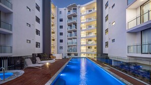A modern building with a swimming pool, multiple balconies, and a wooden deck with lounge chairs. - RIVERVIEWS@The Cosmopolitan Unit 20805 (Sunshine Coast)