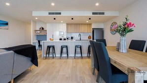 A modern kitchen with a central island, bar stools, and a dining table with chairs. - SALTWATER CALM@ The Cosmopolitan Unit 10508 (Sunshine Coast)