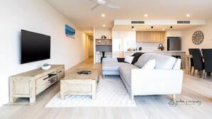A modern living room with a flat-screen TV, a wooden coffee table, a white sofa, and a ceiling fan. - SALTWATER CALM@ The Cosmopolitan Unit 10508 (Sunshine Coast)