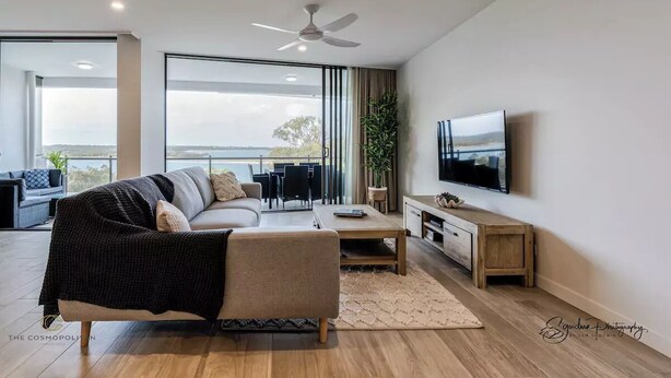 A modern living room with a sofa, coffee table, TV, and a balcony with a view. - SALTWATER CALM@ The Cosmopolitan Unit 10508 (Sunshine Coast)