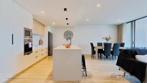 A modern kitchen with a central island, a dining area with a table and chairs, and a large window providing natural light. - SALTWATER CALM@ The Cosmopolitan Unit 10508 (Sunshine Coast)