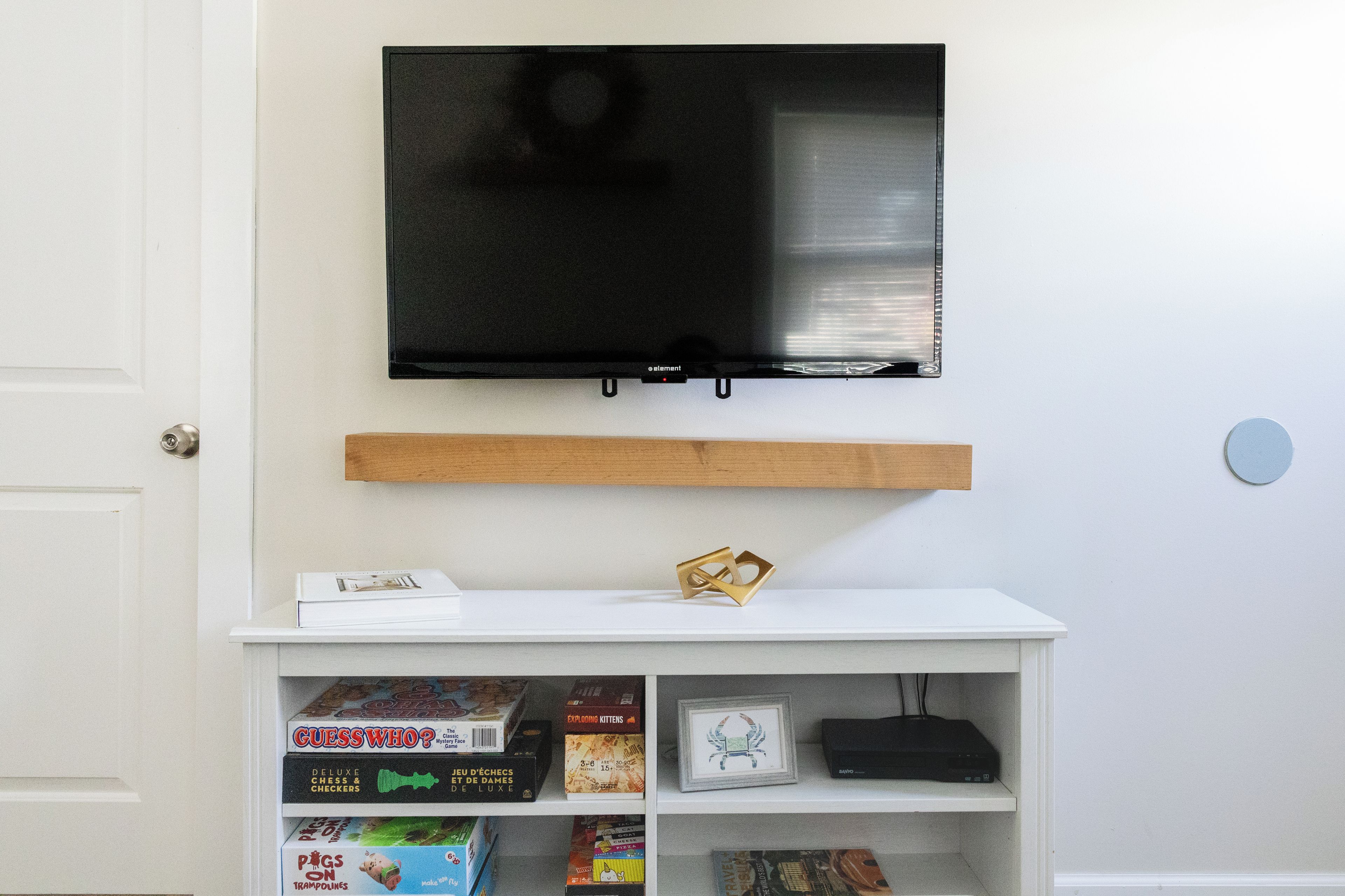 Ein Zimmer mit einem an der Wand befestigten Fernseher über einem weißen Regal, das verschiedene Gesellschaftsspiele und ein gerahmtes Bild beherbergt.