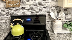 Una cocina con estufa negra, pared de ladrillo, tabla de cortar de madera, planta en maceta y un letrero que dice 'home'.