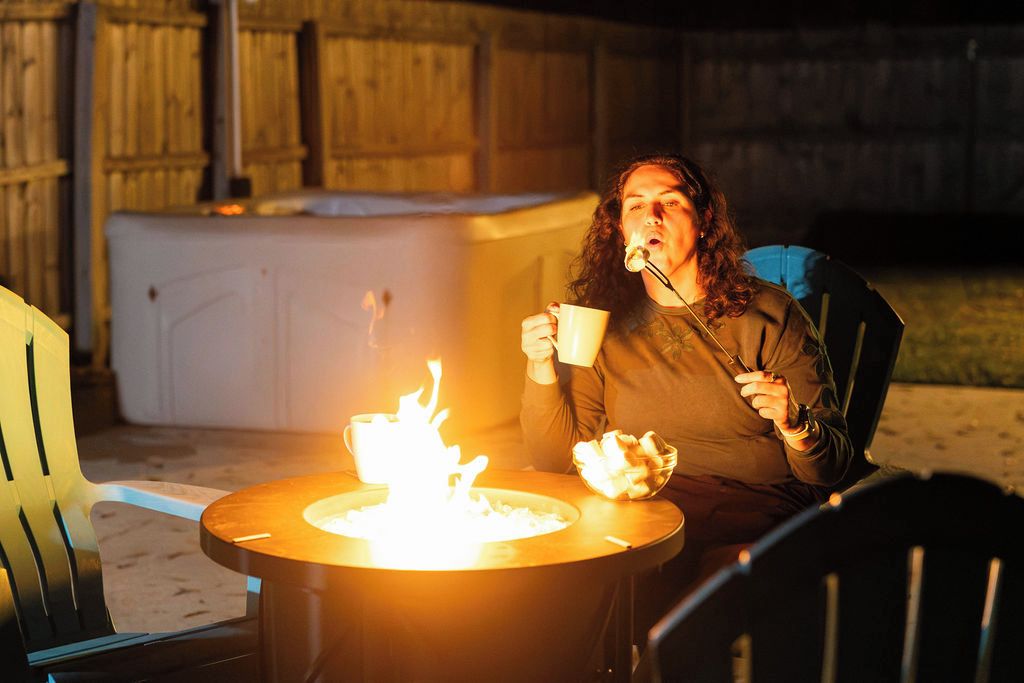A person sitting at a table with a lit fire, holding a cup and a spoon, in an outdoor setting with chairs and a wooden fence in the background.
