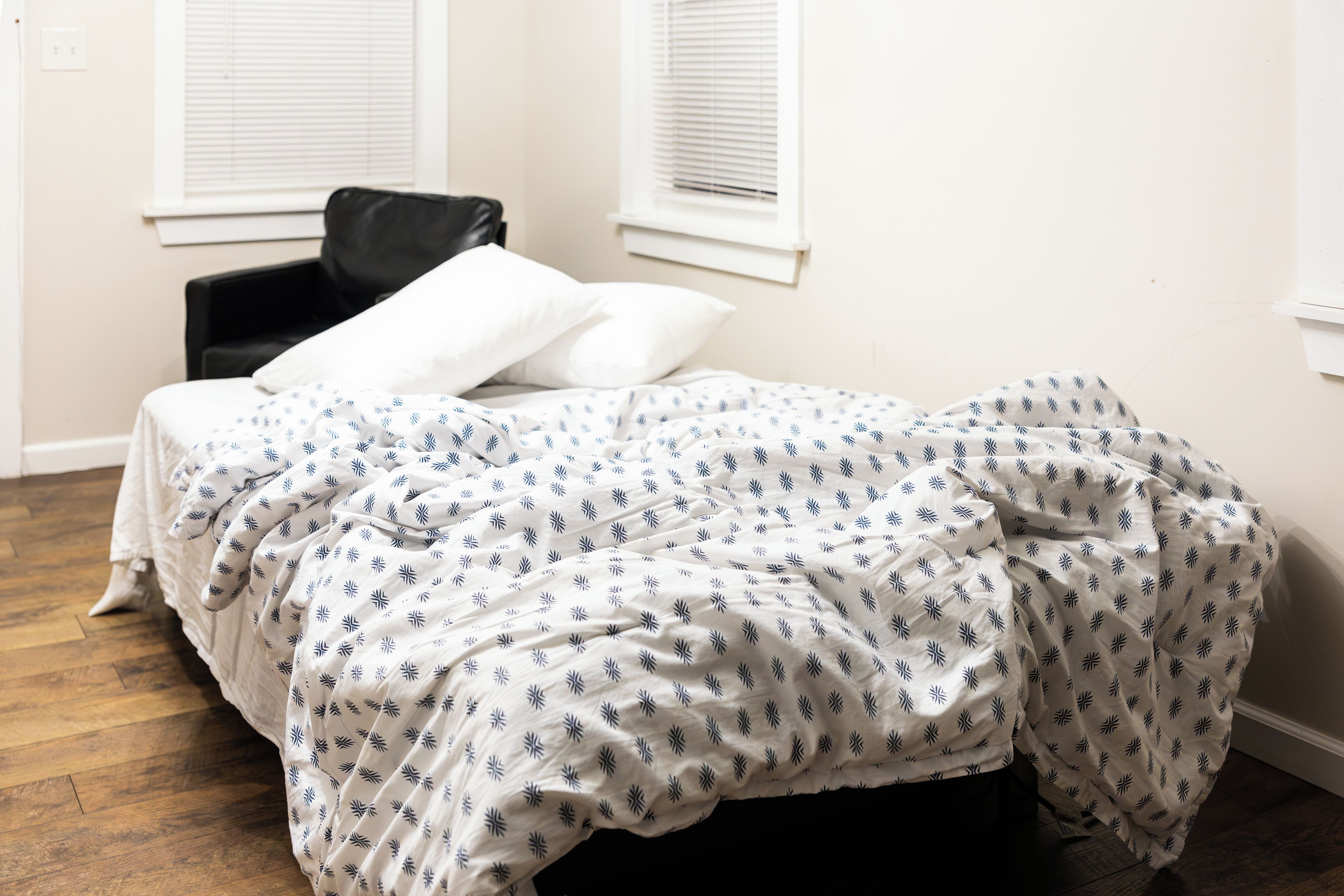 A bedroom with a bed, a chair, and two windows with blinds.