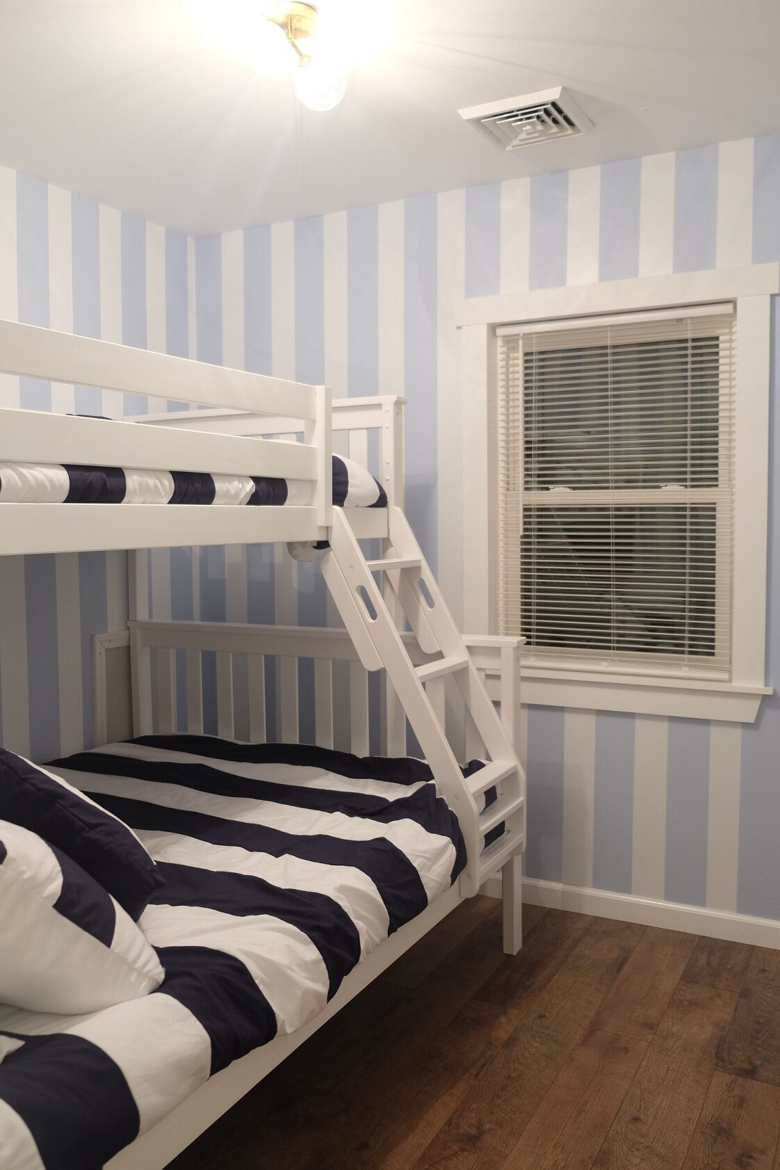 A bunk bed with a striped bedspread, a window with blinds, and a ceiling vent.