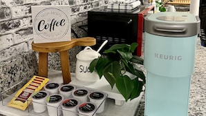Una estación de café con un letrero que dice 'Café recién hecho servido diariamente', una cafetera Keurig y varios tipos de cápsulas de café.